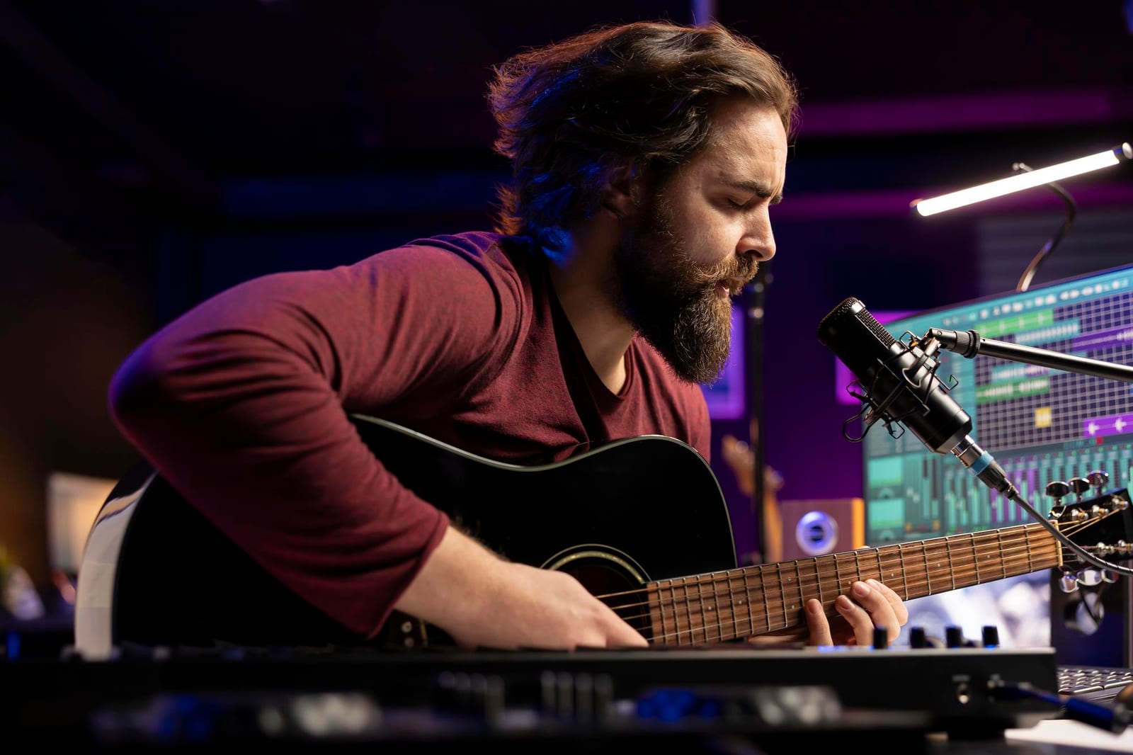 Músico canta e toca violão diante do microfone em um estúdio iluminado em azul e roxo.