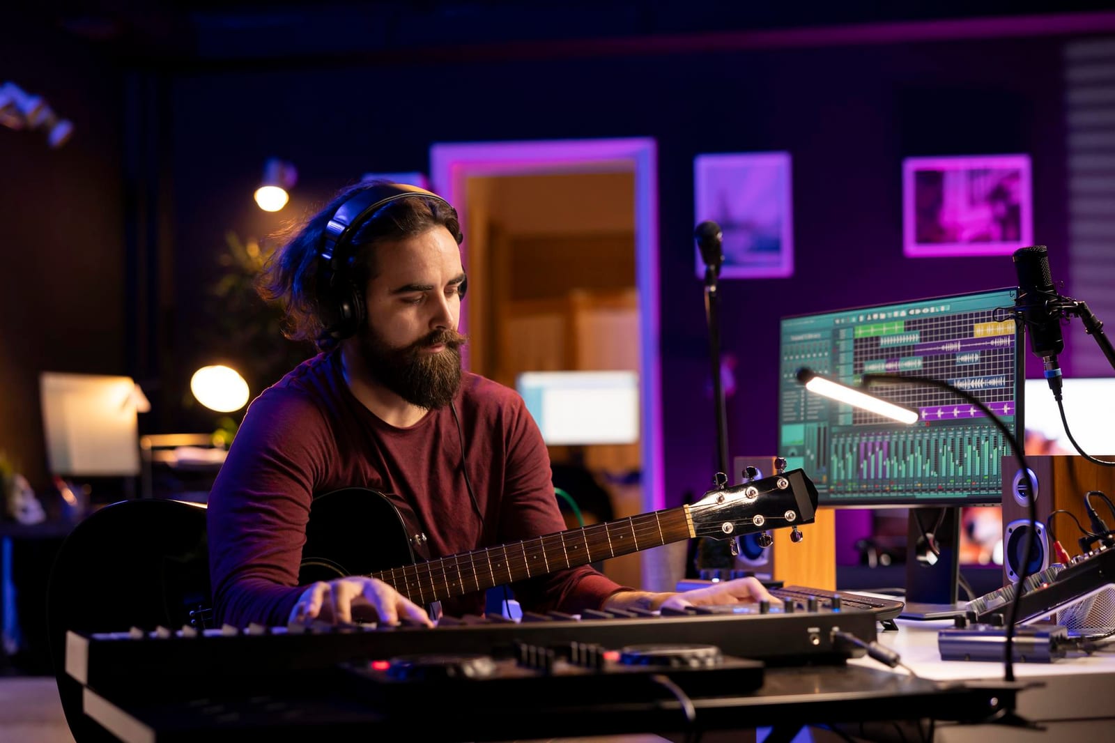 Músico com fones de ouvido toca teclado e violão em um estúdio caseiro iluminado em roxo.