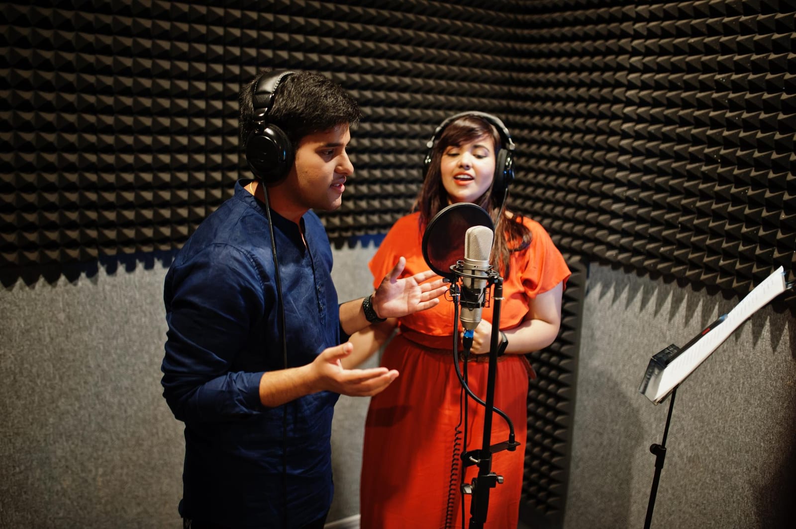 Dupla de cantores grava em uma cabine revestida de espuma acústica com microfone e partitura.